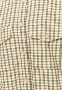 Camicia a quadri verde e beige con due tasche sul petto, chiusura frontale con bottoni e una texture di tessuto leggere. Presenta alette delle tasche arrotondate e bottoni.