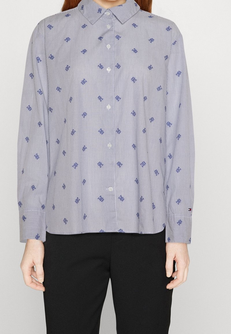 Mujer con camisa celeste de botones con patrones geométricos en azul oscuro y pantalones negros, de pie con los brazos relajados a los lados.