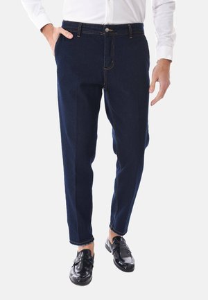 Uomo che indossa jeans slim fit blu scuro, camicia bianca e mocassini in pelle nera, in piedi con una mano in tasca.