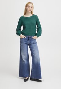 Grüner gestrickter Pullover mit weitem Ausschnitt und langen Ärmeln, kombiniert mit hellblauen weiten Jeans. Schwarze Ballerinas runden das Outfit ab.