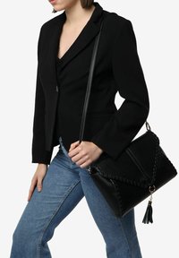 Bolso de mano de cuero negro con forma triangular, detalles trenzados y un adorno de borlas. Se lleva en el hombro con una correa larga.