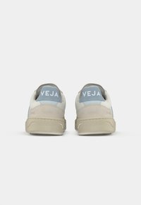 Sneakers med en overdel i beige semsket skinn og lær, lyseblå detaljer og en tan gummisåle. Bak på skoen vises "VEJA"-merket.