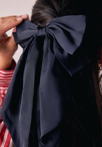 Marineblauer Satin-Haarbogen mit langen, herabhängenden Bändern und glatter Textur, auf dunklem Haar vor hellem Hintergrund befestigt.