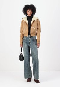 Veste en suede beige courte avec un col en fausse fourrure blanche, associée à un jean en denim bleu à taille haute et coupe ample. Les accessoires comprennent un petit sac à main noir.