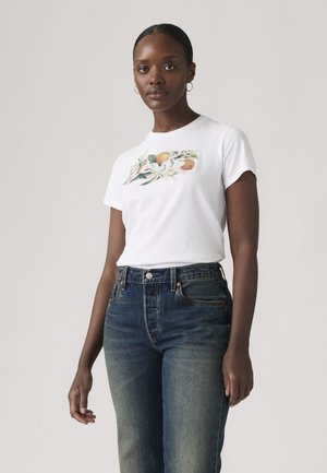 Donna dalla pelle scura e capelli intrecciati che indossa una t-shirt bianca con stampa floreale e jeans blu a vita alta, in piedi davanti a uno sfondo semplice.