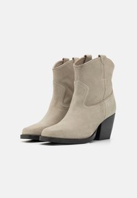 Botas de ante beige hasta el tobillo con puntera afilada, acentos de costura curvada y tacón negro apilado. Lengüetas redondeadas para mayor comodidad al ponérselas.