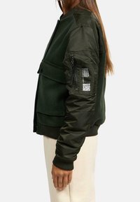 Donkergroene bomberjack met een wollen voorkant, nylon mouwen, geribbelde boorden en een ritszak op de linkerarm met een QR-code.