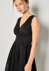 Czarna koronkowa sukienka z dekoltem w serek, bez rękawów, z marszczoną talią i wiązanym detalem. Materiał ma teksturowany wzór kwiatowy.