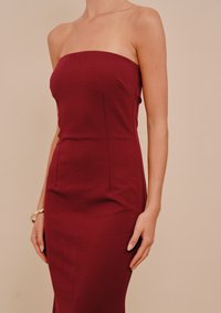 Γυναίκα που φοράει ατσαλάκωτο μπορντό κ fitted φόρεμα χωρίς τιράντες και χρυσό βραχιόλι, στέκεται μπροστά σε απλό μπεζ φόντο.