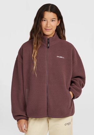 Jonge vrouw met lang haar die een bruine fleece met rits en een beige broek draagt, beide met het O'Neill-logo op een effen achtergrond.