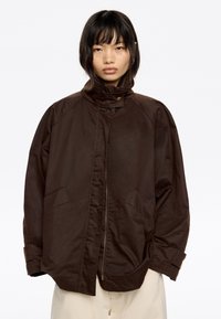 Chaqueta marrón resistente al agua con cuello alto, cierre de cremallera y dos bolsillos laterales. Presenta una textura suave y un diseño de ajuste relajado.