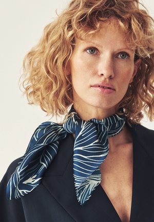 Femme blonde aux cheveux bouclés portant un blazer sombre et une écharpe bleue à motifs de feuilles blanches nouée autour du cou.