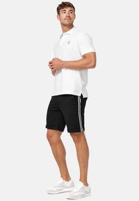 Weißes Polohemd mit Kragen und gesticktem Logo, kombiniert mit schwarzen Shorts mit weißen Seitenstreifen und weißen Sneakern.