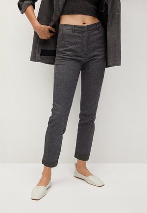 Person trägt graue, strukturierte, zulaufende Hose, graue, gekürzte Jacke, schwarzes, gekürztes Oberteil und weiße, flache Schuhe mit quadratischer Spitze.