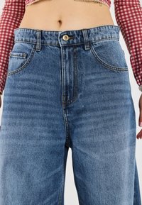 Hochgeschnittene blaue Jeans aus Denim mit geradem Bein, ausgestattet mit zwei vorderen Taschen, einem frontalen Knopfverschluss und kontrastierendem gelbem Nähen.