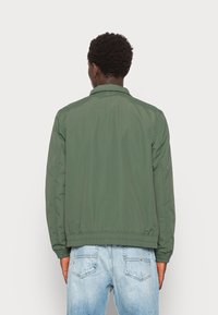 Grön bomberjacka med krage, elastiska mudd, och midjeband, tillverkad av lätt tyg; bärs över ljusblå jeans.