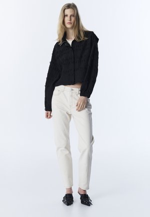 Jonge vrouw staand, gekleed in een gestructureerd zwart cropped jasje, witte jeans met hoge taille en zwarte puntige muiltjes.