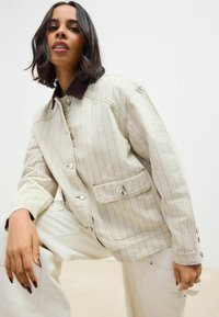 Beige gestreepte jas met een bruine korduroy kraag, voorzien van drukknopen en een klepzak. Model poseert in losse witte broeken.