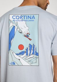 Ανοιχτό μπλε βαμβακερό t-shirt με γραφικό σχέδιο στην πλάτη. Το σχέδιο περιλαμβάνει σκι, βουνά και ένα κόκκινο ήλιο, με μπλε και λευκές λεπτομέρειες.