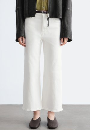 Person trägt weiße, hoch taillierte, weit geschnittene Hosen, schwarzen Gürtel, olivgrünes Hemd mit Knöpfen, schwarze Lederjacke und dunkelbraune Slipper.