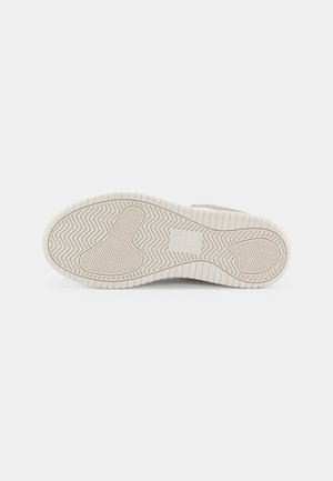 Sålen på en beige sneakers med mønstret slidbane, herunder zigzag og rektangulære grebsområder på en ensartet lys baggrund.