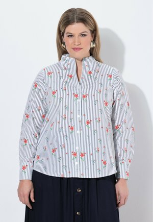 Eine Frau mit glattem blondem Haar trägt eine gestreifte weiße Bluse mit rotem Blumenmuster und große goldene Creolen und steht vor einem weißen Hintergrund.