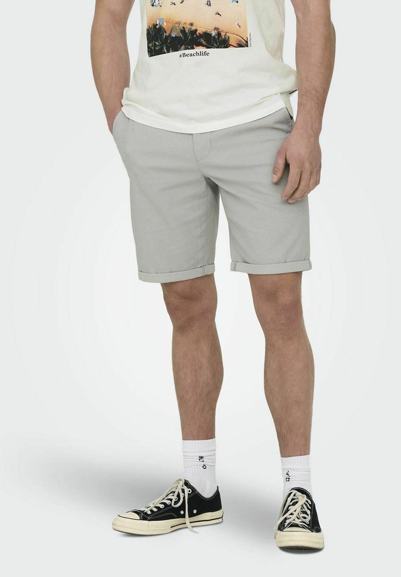 Shorts gris avec une finition texturée, ourlets roulés, associés à un t-shirt graphique et des chaussures noires. Les shorts ont une coupe relaxée et un style décontracté.