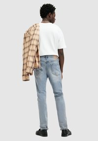 Jeans de color azul claro con un corte recto, combinados con una camiseta blanca. Una camisa de cuadros beige colgada sobre un hombro. Zapatos negros.