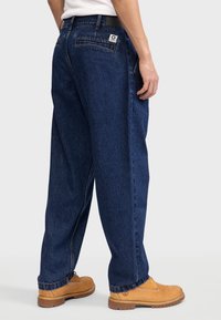 Jean en denim bleu foncé avec une coupe décontractée, poches arrière visibles et un patch logo. Assortis à des bottes de travail couleur sable.