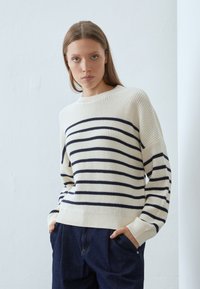 Stripet genser i kremfarge og marineblå, strikket tekstur, rund hals, senkede skuldre, ribbestrikkede mansjetter og fald, sammen med mørke jeans.