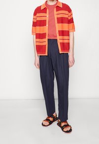 Kortärmad röd- och orange-randig skjorta, enfärgad orange skjorta under, marinblå löst sittande byxor, svarta sandaler med orange detaljer.