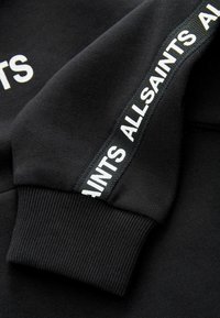 Sweat-shirt noir avec poignets côtelés, arborant un logo blanc "ALLSAINTS" le long de la manche dans un design en bande contrastante. Texture lisse.