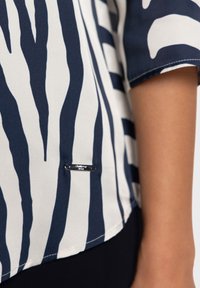 Blouse bleu marine et blanche avec un motif à rayures zébrées, tissu léger et petit logo métallique près de l'ourlet.