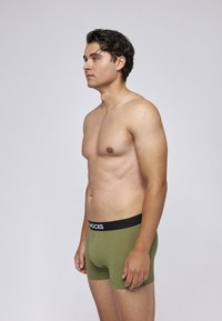 Grüne Baumwoll-Boxershorts mit einem schwarzen Bund, auf dem "KNOCKS" steht. Das Modell wird im Profil vor einem neutralen Hintergrund gezeigt.