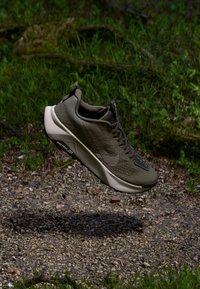Dunkelgrüner Trailrunningschuh mit weißer Sohle schwebt über einem Kiesweg, der von grünem Gras und Laub umgeben ist.