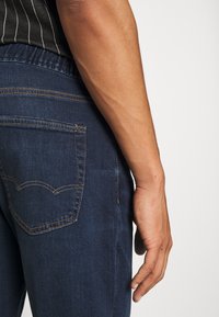 Bras droit et main reposant à côté de la hanche d'une personne portant un jean bleu foncé et un haut noir à rayures blanches.