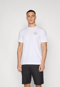 Weißes Baumwoll-T-Shirt mit kurzen Ärmeln, das ein rundes Logo auf der Brust zeigt. Kombiniert mit schwarzen Shorts für einen Kontrast.