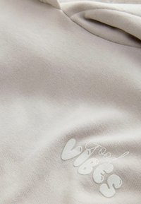 Ljust beige hoodie med en mjuk textur; har en upphöjd vit "Good Vibes" grafik på framsidan, vilket betonar en avslappnad design.
