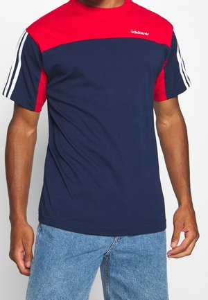 Man wearing a navy blue and red Adidas t-shirt with white stripes on sleeves and light blue denim jeans, standing against a white background.