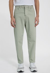 Pantalones de color verde claro, hechos de algodón, con un corte relajado, bolsillos frontales y cremallera en la bragueta. Combinados con zapatillas blancas y grises.