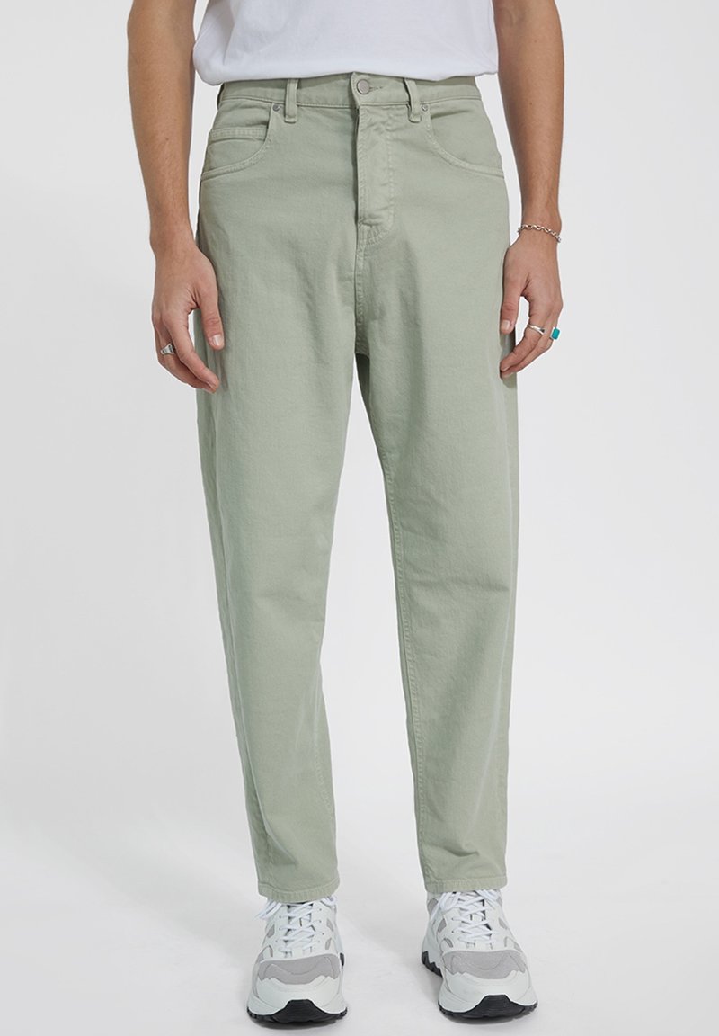 Pantalones de color verde claro, hechos de algodón, con un corte relajado, bolsillos frontales y cremallera en la bragueta. Combinados con zapatillas blancas y grises.