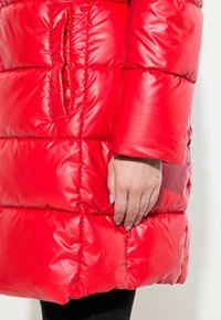 Abrigo acolchado rojo con tela brillante, que presenta secciones acolchadas, un bolsillo lateral y una silueta relajada. Las mangas terminan en la muñeca.