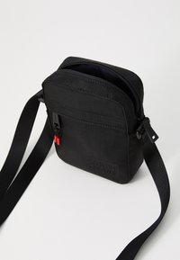 Čierna kompaktná crossbody taška s nastaviteľným popruhom, predná zipsová kapsa s červenými ťahadlami a embosovaným logom "JOOP! JEANS".