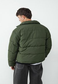 Grüne Pufferjacke mit Kragen, bestehend aus horizontal gesteppter Struktur und lockerer Passform. Kombiniert mit weiten grauen Hosen.