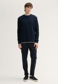 Maglione a maniche lunghe navy a coste con scollo rotondo, indossato sopra una camicia bianca, abbinato a pantaloni scuri e sneakers nere.