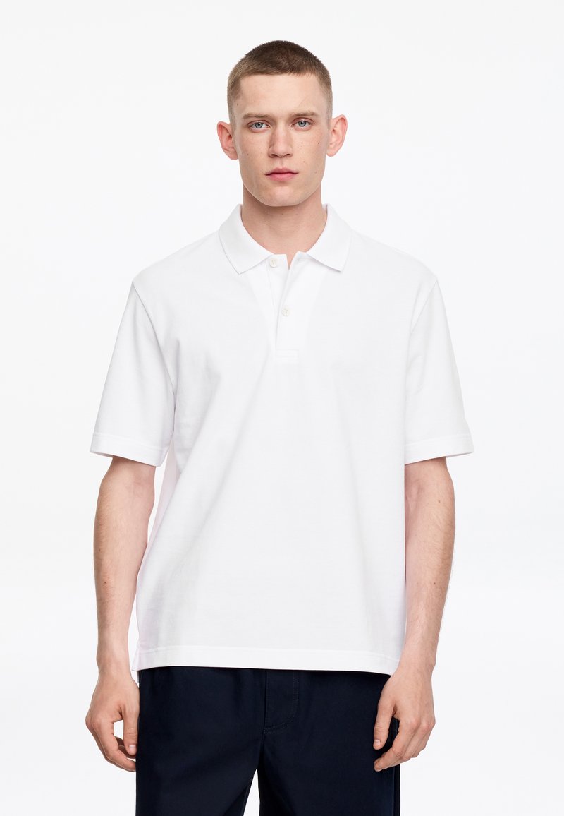 Jeune homme aux cheveux courts portant un polo blanc uni à manches courtes et un pantalon foncé, debout devant un fond blanc.