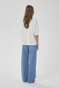 Ženska z dolgimi rjavimi lasmi nosi belo bluzo s širokimi rokavi in široke modre kavbojke, stoji obrnjena stran proti enostavni sivi ozadju.