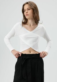 Wit geribd shirt met lange mouwen, een v-hals en een overslag aan de voorkant, gecombineerd met zwarte broek met hoge taille en een tailleband met strik.