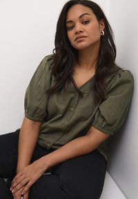 Blouse vert olive avec un col en V, des manches bouffantes et des boutons dorés. Modèle portant un pantalon noir, assis contre un fond clair.