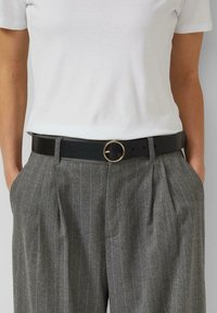 Zwarte leren riem met een ronde gouden gesp. Draagt bij een wit T-shirt en grijze broek met krijtstrepen. Heeft een gladde textuur.